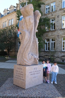 A stone memorial monument is inscribed with text in different languages, located in a public square. Three children are standing beside it, two girls and a boy, smiling. The building in the background is made of stone with multiple windows, suggesting a historical or official setting. There are also flags visible behind the monument.