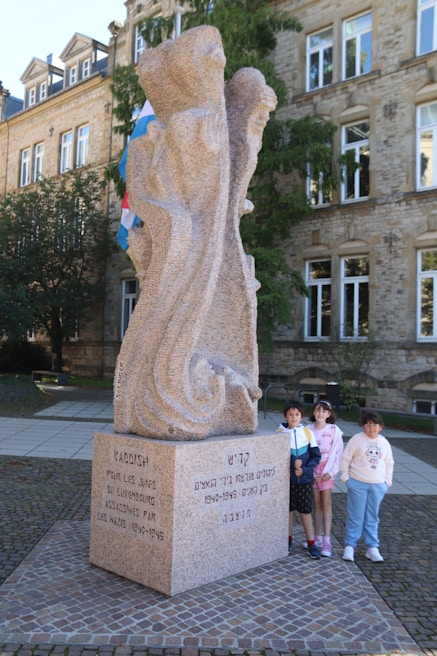 A stone memorial monument is inscribed with text in different languages, located in a public square. Three children are standing beside it, two girls and a boy, smiling. The building in the background is made of stone with multiple windows, suggesting a historical or official setting. There are also flags visible behind the monument.