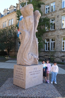 A stone memorial monument is inscribed with text in different languages, located in a public square. Three children are standing beside it, two girls and a boy, smiling. The building in the background is made of stone with multiple windows, suggesting a historical or official setting. There are also flags visible behind the monument.