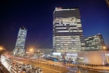 This is a nighttime cityscape featuring several high-rise buildings. The tallest building has many illuminated windows and a sign that reads 'Vodafone' at the top. A second round-shaped building is slightly tilted to the left. There are several cars on the road, indicating heavy traffic, and a few streetlights provide additional light to the scene. The lower part of the image shows a commercial establishment labeled 'Mega Image' surrounded by people and more cars.