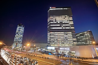 This is a nighttime cityscape featuring several high-rise buildings. The tallest building has many illuminated windows and a sign that reads 'Vodafone' at the top. A second round-shaped building is slightly tilted to the left. There are several cars on the road, indicating heavy traffic, and a few streetlights provide additional light to the scene. The lower part of the image shows a commercial establishment labeled 'Mega Image' surrounded by people and more cars.