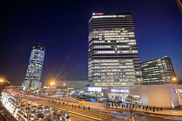 This is a nighttime cityscape featuring several high-rise buildings. The tallest building has many illuminated windows and a sign that reads 'Vodafone' at the top. A second round-shaped building is slightly tilted to the left. There are several cars on the road, indicating heavy traffic, and a few streetlights provide additional light to the scene. The lower part of the image shows a commercial establishment labeled 'Mega Image' surrounded by people and more cars.