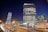 This is a nighttime cityscape featuring several high-rise buildings. The tallest building has many illuminated windows and a sign that reads 'Vodafone' at the top. A second round-shaped building is slightly tilted to the left. There are several cars on the road, indicating heavy traffic, and a few streetlights provide additional light to the scene. The lower part of the image shows a commercial establishment labeled 'Mega Image' surrounded by people and more cars.