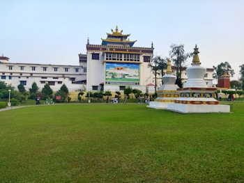 A large, multi-story building with traditional architecture, featuring a colorful mural on the front. In the foreground, there are two white stupa structures with red and gold accents. The building is surrounded by a well-maintained green lawn with trees and bushes, and several people are walking around.