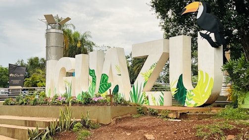 Large white letters spelling 'IGUAZU' are decorated with vibrant, tropical leaf designs. A toucan is perched on the letter 'U'. Flowers and greenery surround the base, with a background of palm trees and a tall metallic structure.