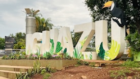 Large white letters spelling 'IGUAZU' are decorated with vibrant, tropical leaf designs. A toucan is perched on the letter 'U'. Flowers and greenery surround the base, with a background of palm trees and a tall metallic structure.