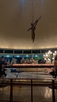 A dimly lit interior with a large crucifix hanging from the ceiling. The foreground has a row of lit candles and flower arrangements on a table. People are seated on benches facing the candles. A chandelier and religious statues are visible in the background.