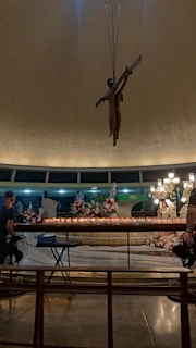 A dimly lit interior with a large crucifix hanging from the ceiling. The foreground has a row of lit candles and flower arrangements on a table. People are seated on benches facing the candles. A chandelier and religious statues are visible in the background.