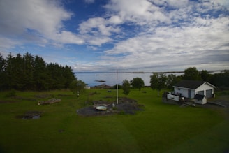A peaceful landscape featuring a grassy field with a small house and garage. There is a Norwegian flag on a pole in the middle of the lawn. The scene is completed by a backdrop of trees on the left and a calm body of water on the right, under a sky with clouds and patches of blue.