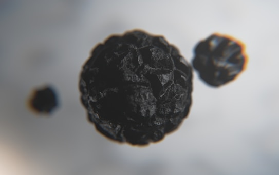 Close-up view of three black peppercorns against a blurred, light grey background. The image captures the textured surface of the peppercorns, allowing their rough exterior and spherical shape to stand out prominently.