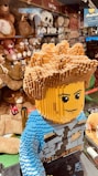 A large LEGO model of a person with detailed hair and facial features stands in the foreground. The model is wearing a blue outfit with blocky design elements. In the background, shelves are lined with a variety of plush toys, including teddy bears and other stuffed animals, with signs indicating 'Soft Toys.' The setting appears to be a toy store.