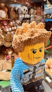 A large LEGO model of a person with detailed hair and facial features stands in the foreground. The model is wearing a blue outfit with blocky design elements. In the background, shelves are lined with a variety of plush toys, including teddy bears and other stuffed animals, with signs indicating 'Soft Toys.' The setting appears to be a toy store.