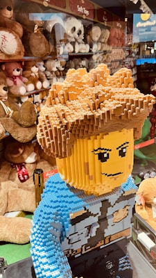 A large LEGO model of a person with detailed hair and facial features stands in the foreground. The model is wearing a blue outfit with blocky design elements. In the background, shelves are lined with a variety of plush toys, including teddy bears and other stuffed animals, with signs indicating 'Soft Toys.' The setting appears to be a toy store.