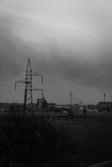 A black-and-white image featuring several electrical transmission towers standing prominently across a flat landscape. In the background, there are small buildings and structures, partially obscured by the haze. The foreground includes bushy vegetation and an expansive open field. The sky is overcast, adding to the moody atmosphere.