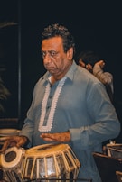 A man in a blue traditional outfit plays a tabla, a pair of Indian percussion instruments. He appears focused and is captured in a dimly lit setting, which adds depth to the scene.
