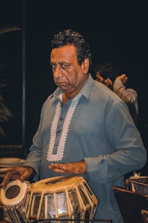 A man in a blue traditional outfit plays a tabla, a pair of Indian percussion instruments. He appears focused and is captured in a dimly lit setting, which adds depth to the scene.