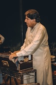 A man wearing a traditional shalwar kameez is playing a harmonium. He is focused and standing beside the musical instrument. The background is dark, emphasizing the subject and the harmonium.