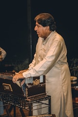A man wearing a traditional shalwar kameez is playing a harmonium. He is focused and standing beside the musical instrument. The background is dark, emphasizing the subject and the harmonium.