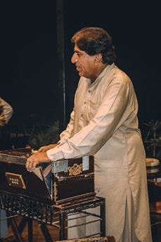 A man wearing a traditional shalwar kameez is playing a harmonium. He is focused and standing beside the musical instrument. The background is dark, emphasizing the subject and the harmonium.