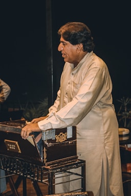 A man wearing a traditional shalwar kameez is playing a harmonium. He is focused and standing beside the musical instrument. The background is dark, emphasizing the subject and the harmonium.