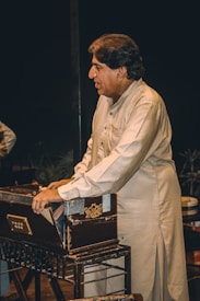 A man wearing a traditional shalwar kameez is playing a harmonium. He is focused and standing beside the musical instrument. The background is dark, emphasizing the subject and the harmonium.