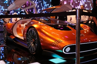 A futuristic orange concept car with gull-wing doors open is displayed indoors. The interior includes quilted seating and an illuminated dashboard. The car is surrounded by people taking pictures and admiring it in a dimly lit space with a pixelated digital backdrop.