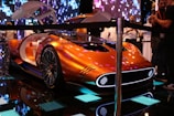 A futuristic concept car with a sleek metallic orange design, featuring a streamlined body and large illuminated wheels. The car door is open, revealing a luxurious white and orange interior. The setting is an indoor exhibit with digital lighting effects and a crowd of people observing the car.