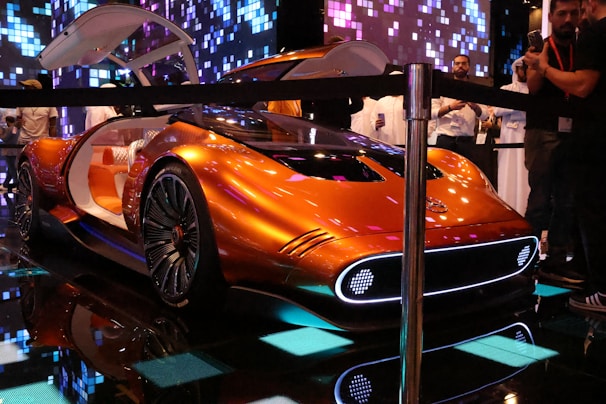A futuristic concept car with a sleek metallic orange design, featuring a streamlined body and large illuminated wheels. The car door is open, revealing a luxurious white and orange interior. The setting is an indoor exhibit with digital lighting effects and a crowd of people observing the car.