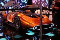 A futuristic concept car with a sleek metallic orange design, featuring a streamlined body and large illuminated wheels. The car door is open, revealing a luxurious white and orange interior. The setting is an indoor exhibit with digital lighting effects and a crowd of people observing the car.