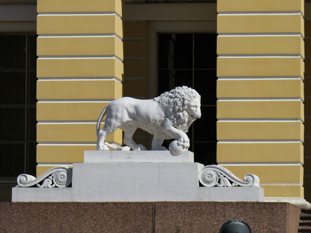 A white lion statue positioned atop a pedestal with ornate decorations. The lion is depicted in a standing pose with one paw resting on a spherical object. The background features a building facade with yellow and cream-colored horizontal stripes.