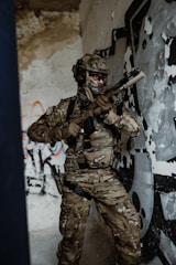 A soldier in full camouflage gear is holding a rifle inside a room with peeling paint and graffiti on the walls. The soldier is wearing tactical gear and a helmet, appearing alert and ready for action.
