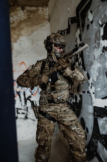 A soldier in full camouflage gear is holding a rifle inside a room with peeling paint and graffiti on the walls. The soldier is wearing tactical gear and a helmet, appearing alert and ready for action.