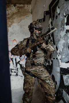 A soldier in full camouflage gear is holding a rifle inside a room with peeling paint and graffiti on the walls. The soldier is wearing tactical gear and a helmet, appearing alert and ready for action.