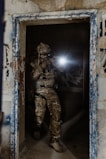A person in military gear is holding a firearm and shining a tactical flashlight in a dimly lit, decaying urban environment, standing in a doorway of an abandoned building with graffiti on the walls.