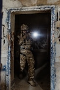 A person in military gear is holding a firearm and shining a tactical flashlight in a dimly lit, decaying urban environment, standing in a doorway of an abandoned building with graffiti on the walls.