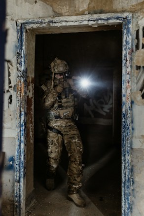 A person in military gear is holding a firearm and shining a tactical flashlight in a dimly lit, decaying urban environment, standing in a doorway of an abandoned building with graffiti on the walls.
