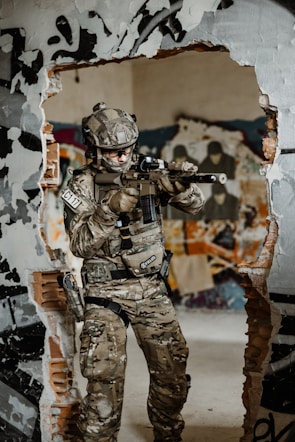 A person in military camouflage gear and tactical equipment is holding a rifle. The individual stands in front of a wall with a large hole, surrounded by graffiti and posters. The setting appears to be an abandoned or urban area with exposed bricks and a rough, rugged environment.