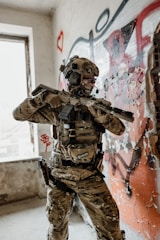 A person in military-style gear is positioned in a room with peeling paint and graffiti on the walls. They are holding a rifle and wearing camouflage clothing, a helmet with a headset, and protective eyewear. The environment appears to be an abandoned or urban setting, with visible graffiti including hearts and abstract shapes.