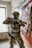 A person in military-style gear is positioned in a room with peeling paint and graffiti on the walls. They are holding a rifle and wearing camouflage clothing, a helmet with a headset, and protective eyewear. The environment appears to be an abandoned or urban setting, with visible graffiti including hearts and abstract shapes.