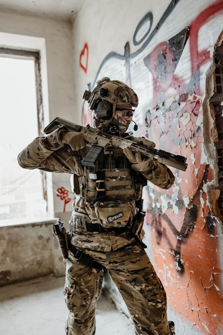 A person in military-style gear is positioned in a room with peeling paint and graffiti on the walls. They are holding a rifle and wearing camouflage clothing, a helmet with a headset, and protective eyewear. The environment appears to be an abandoned or urban setting, with visible graffiti including hearts and abstract shapes.