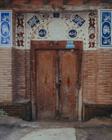 A rustic, double wooden door set within a decorative brick archway. Arabic calligraphy and ornamental patterns adorn the arch, incorporating intricate designs with blue and white tiles. The wood appears weathered, adding a historic feel to the architectural setting, while bricks add texture and depth.