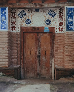 A rustic, double wooden door set within a decorative brick archway. Arabic calligraphy and ornamental patterns adorn the arch, incorporating intricate designs with blue and white tiles. The wood appears weathered, adding a historic feel to the architectural setting, while bricks add texture and depth.