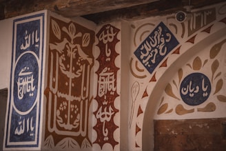 Traditional Arabic calligraphy is elaborately displayed on wall panels, featuring intricate designs and patterns in contrasting colors such as blue, brown, and white. The text appears to be religious or cultural, set against a textured background with decorative motifs.