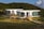 Three modern, oval-shaped, modular white houses are situated in a lush, green hilly landscape. Each house has large glass windows and wooden staircases leading up to their entrances. A beige canopy tent is set up in the grassy area in front of the houses, and a person is seen walking near the structure. The sun casts warm light, enhancing the scenic and serene atmosphere.
