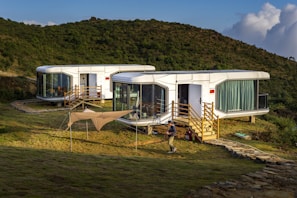 Three modern, oval-shaped, modular white houses are situated in a lush, green hilly landscape. Each house has large glass windows and wooden staircases leading up to their entrances. A beige canopy tent is set up in the grassy area in front of the houses, and a person is seen walking near the structure. The sun casts warm light, enhancing the scenic and serene atmosphere.