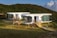 Three modern, oval-shaped, modular white houses are situated in a lush, green hilly landscape. Each house has large glass windows and wooden staircases leading up to their entrances. A beige canopy tent is set up in the grassy area in front of the houses, and a person is seen walking near the structure. The sun casts warm light, enhancing the scenic and serene atmosphere.