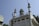 A white mosque with green and turquoise accents features several ornate domes and tall, slender minarets with decorative elements. There are multiple arched windows, and a speaker is visible near one of the domes. The structure is surrounded by trees, and power lines run across the scene.