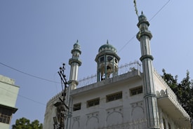 A white mosque with green and turquoise accents features several ornate domes and tall, slender minarets with decorative elements. There are multiple arched windows, and a speaker is visible near one of the domes. The structure is surrounded by trees, and power lines run across the scene.