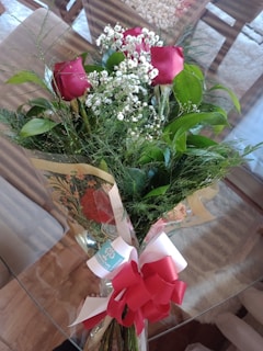 A bouquet of red roses and small white flowers with lush green foliage, wrapped in decorative paper and fastened with a large red and white bow.