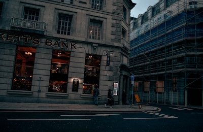 A street scene featuring a corner building with large windows, labeled as 'Kett's Bank.' The bank's windows reveal a warmly lit interior with decorative elements. Adjacent buildings are covered in scaffolding, suggesting construction or renovation work. A few pedestrians are visible on the sidewalk, and a signpost is placed near the corner.